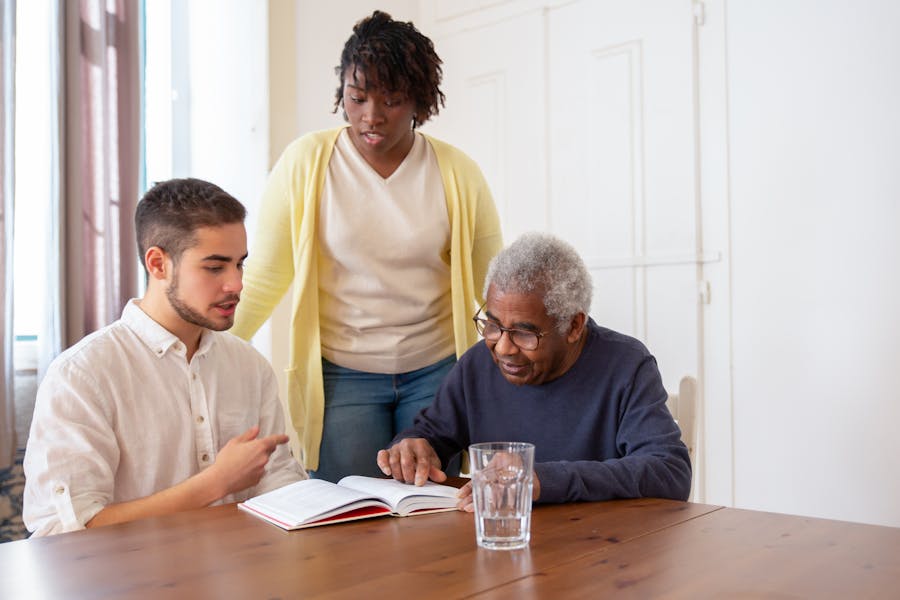 A young carer reads with an older man at a table while a colleague supervises